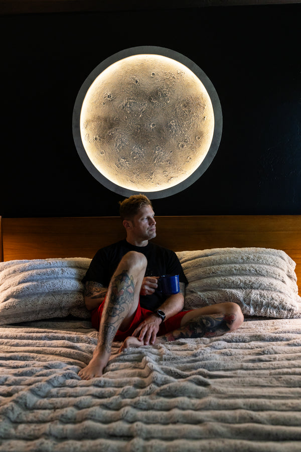 Person sitting on a bed in early morning with a large moon light above them.