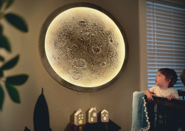 Child sitting in a crib in front of a large moon-shaped light fixture on a wall.